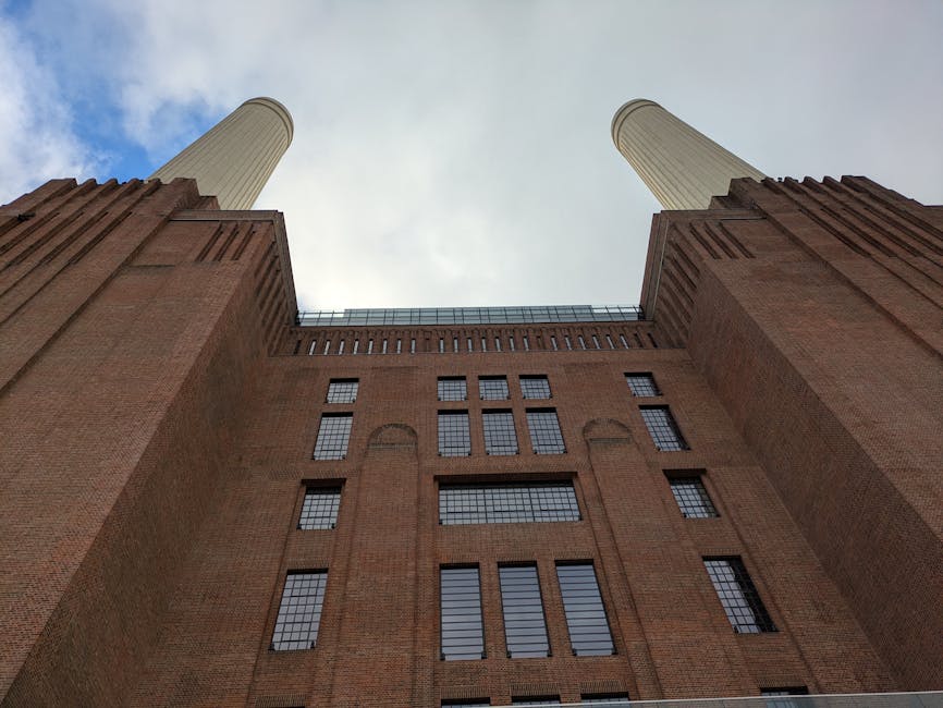 A large historic power station building situated along a riverbank, constructed from red brick with a textured surface. The structure features four tall, cream-colored smokestacks extending vertically